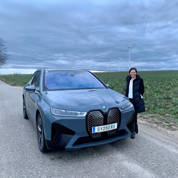 Eine Frau steht neben einem grauen BMW iX vor einem Feld mit Windrädern im Hintergrund.