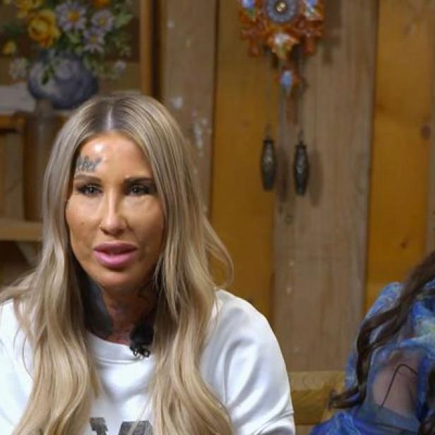 Zwei Frauen sitzen vor einer hölzernen Wand, eine mit einer Tiara.