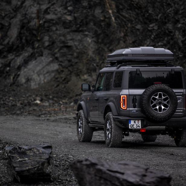 Ein grauer Ford Bronco mit Dachzelt fährt auf einer Schotterstraße.