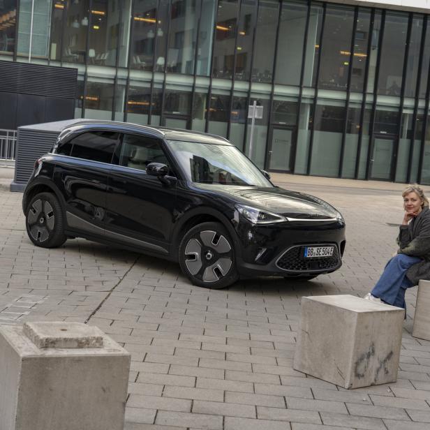 Eine Frau sitzt neben einem schwarzen Smart Elektroauto auf Betonblöcken.