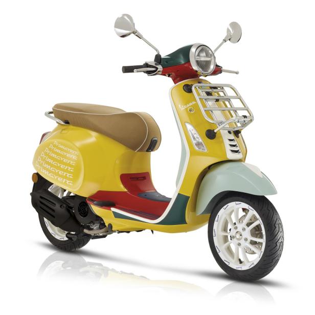 Eine gelbe Vespa Primavera mit farbigen Akzenten steht auf einem weißen Hintergrund.