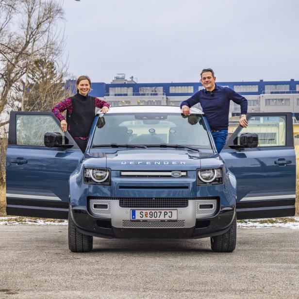 Ein blauer Land Rover Defender mit geöffneten Türen und zwei Personen, die sich darauf abstützen.